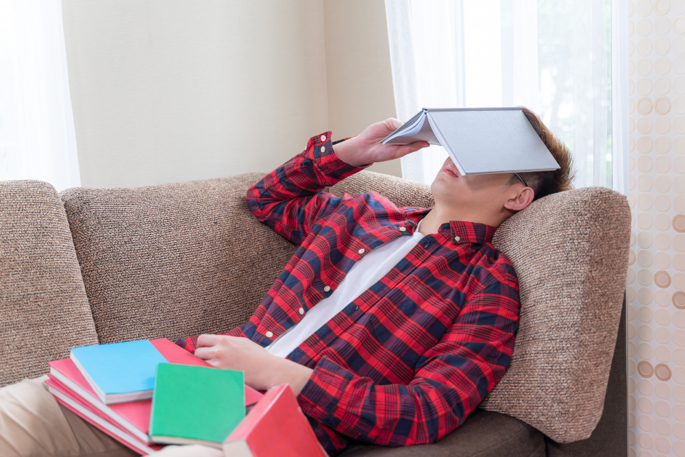 Joven cansado y frustrado en el sofá rodeado de libros, simbolizando el agotamiento por estudiar sin gestionar su tiempo.